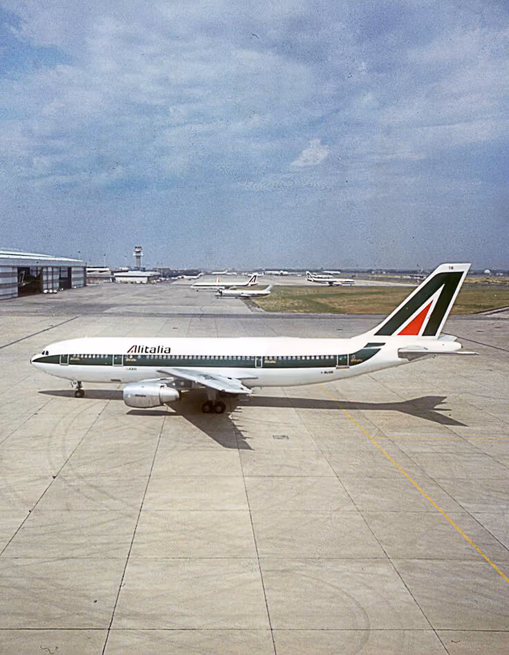 Alitalia airplane parked on an airport tarmac with hangars and other planes in background.
