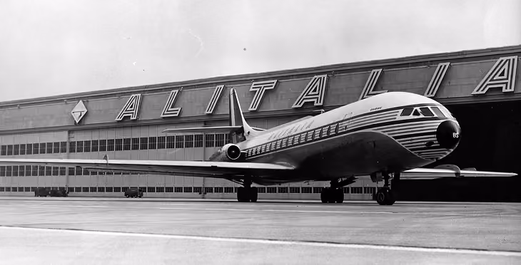 Black and white photo of a vintage airplane parked in front of a building with large ALITALIA letters.
