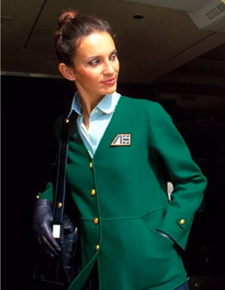Woman in green jacket with gold buttons and black gloves looking to the side in low light.